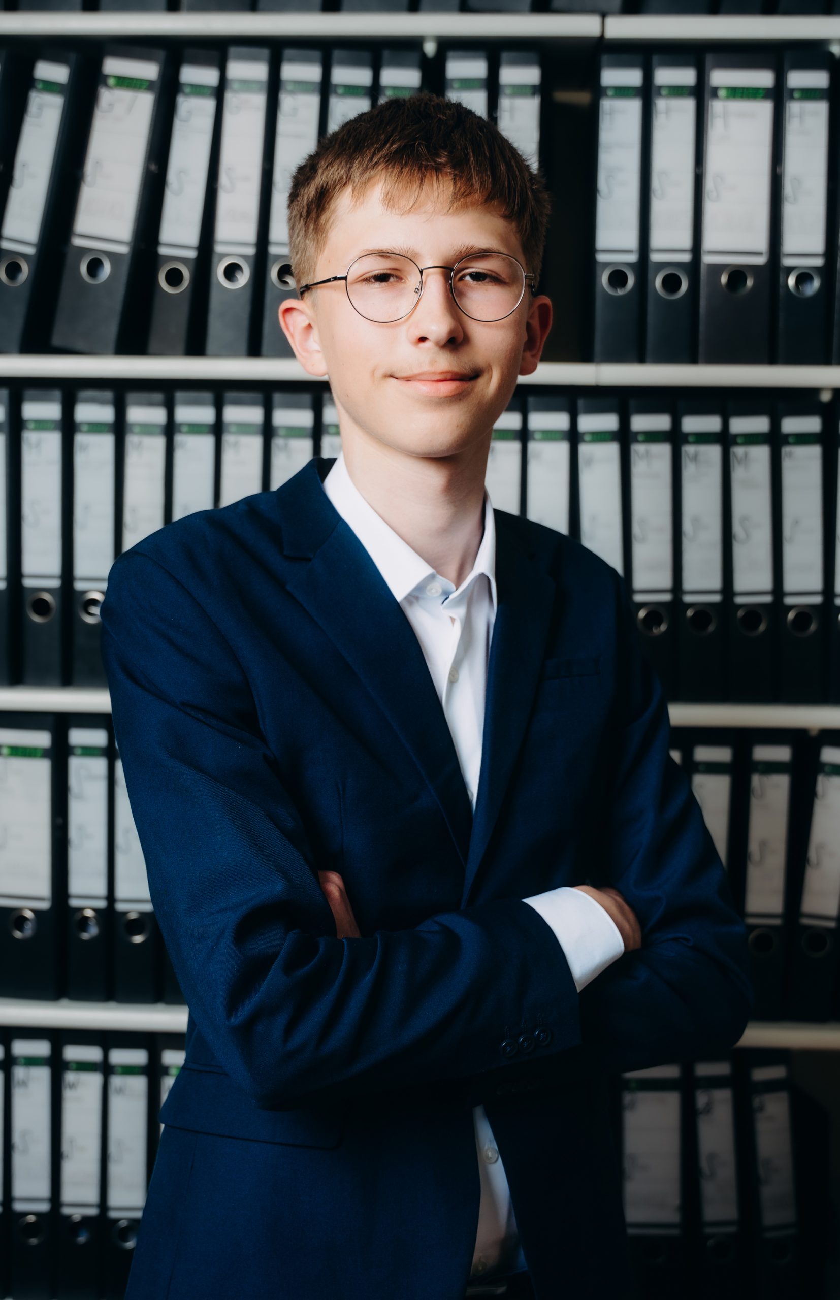 Ein junger Auszubildender mit Brille und blauem Anzug steht lächelnd 